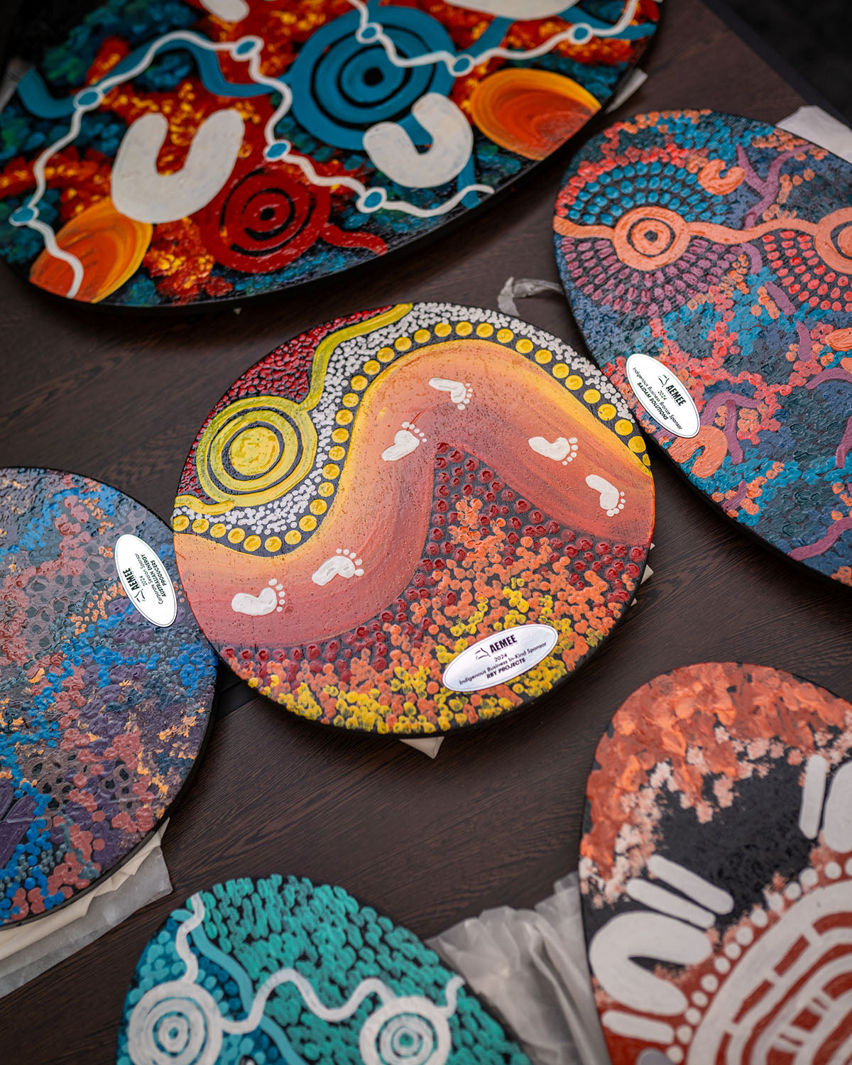 An angled close-up view of multiple custom wooden award plaques displaying vibrant yellow, orange, and pink traditional First Nations artwork on a display table.