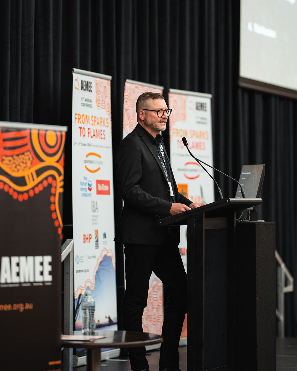 Keynote speaker Kent Matla presenting at the main stage podium under corporate partner banners at the annual national AEMEE conference.