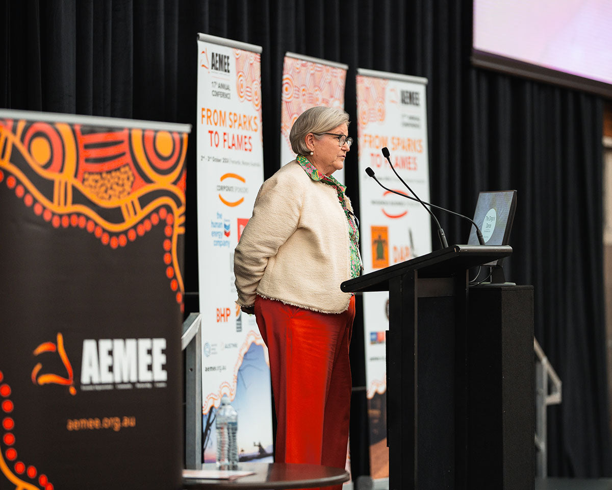 Amanda Healy of Warrikal, wearing red trousers and a cream jacket, presenting "The Dingo's Dance - the Warrikal Story" from the stage podium at the annual AEMEE conference.