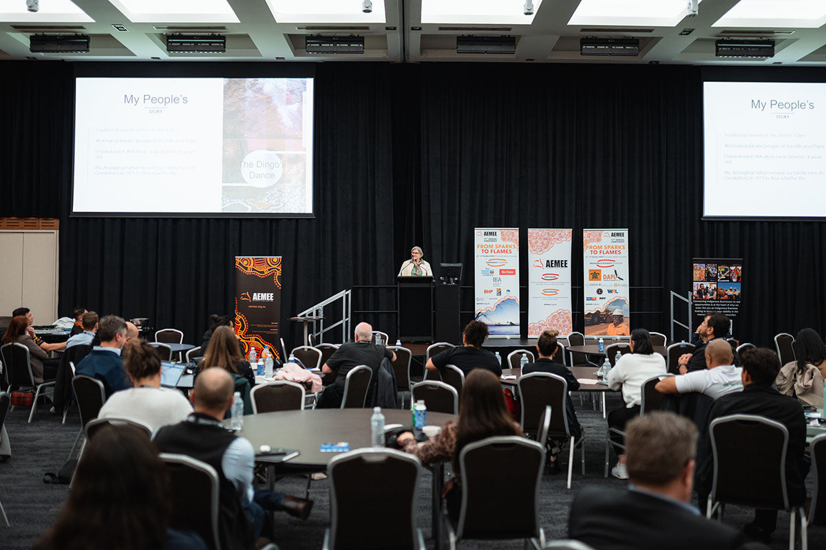 A wide perspective of the packed conference hall watching Amanda Healy's presentation with overhead screens displaying "My People's" and "The Dingo's Dance" slide text.