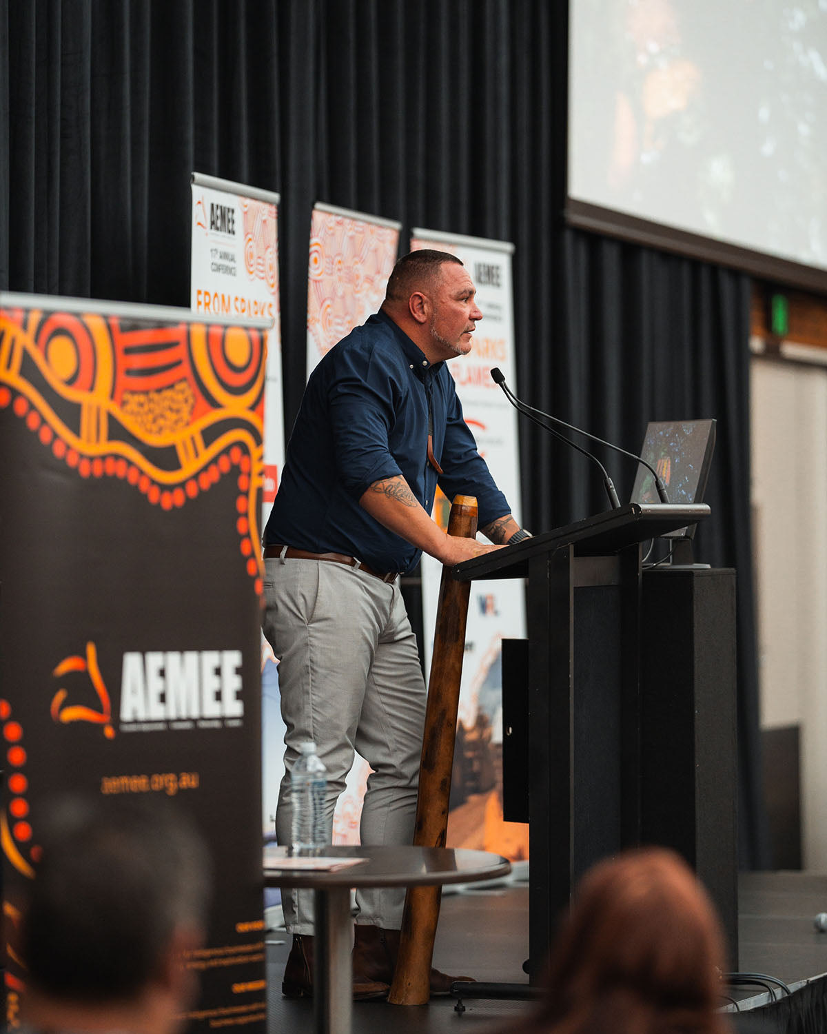 A male cultural presenter standing at the stage podium microphone with a yidaki resting beside him during the closing plenary session at the AEMEE conference.