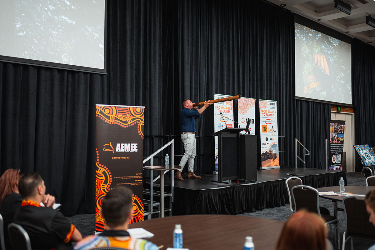A wide shot of a performer playing a traditional wooden didgeridoo on the main stage to mark the closing ceremony of the national AEMEE conference.