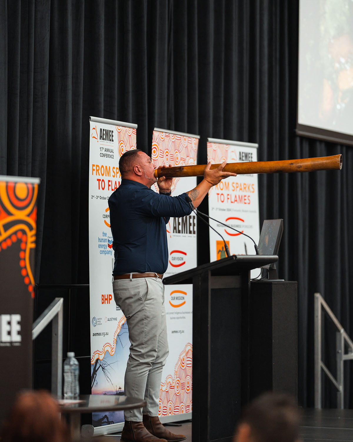 A close-up view of a First Nations man playing a long wooden didgeridoo on stage in front of "From Sparks to Flames" banners at the annual national AEMEE conference.