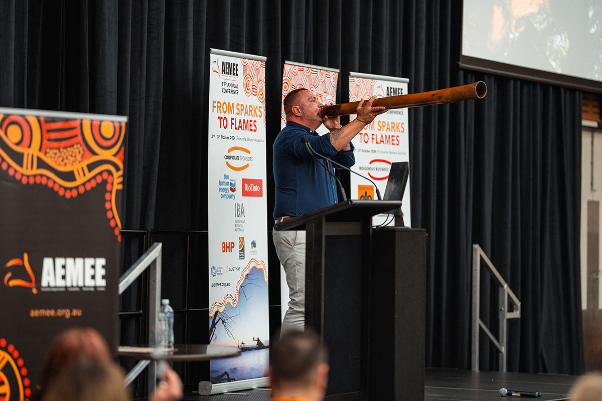 A side profile shot of a male First Nations performer playing a long wooden didgeridoo on stage behind the podium at the annual AEMEE conference.
