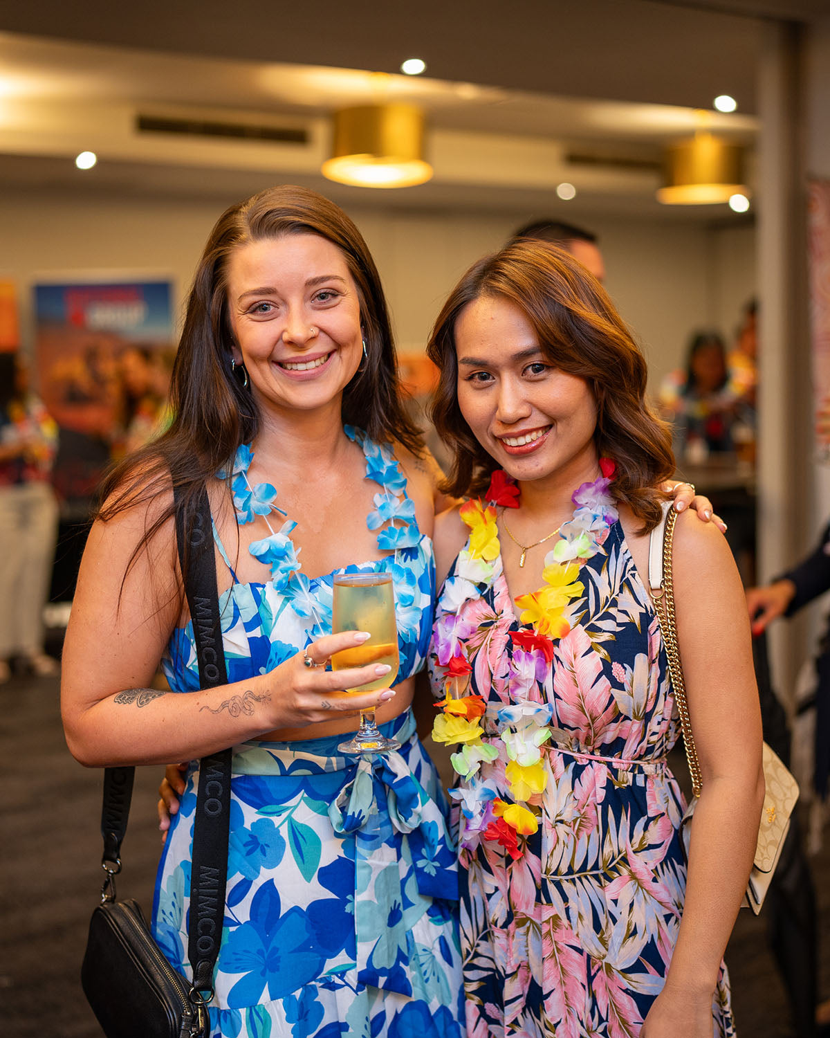 Two female attendees in matching blue, pink, and white floral dresses smiling with drinks during the themed corporate networking evening.