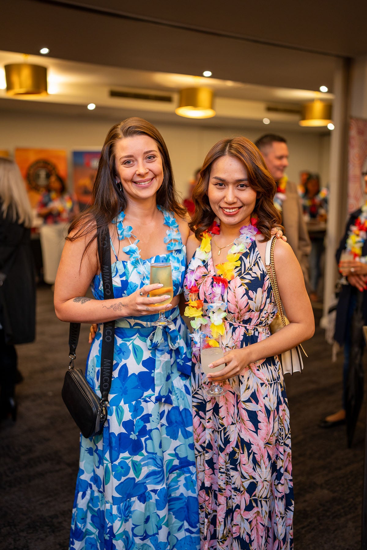 Two women enjoying drinks and showcasing bright floral print outfits and flower leis during a festive breakout social session.