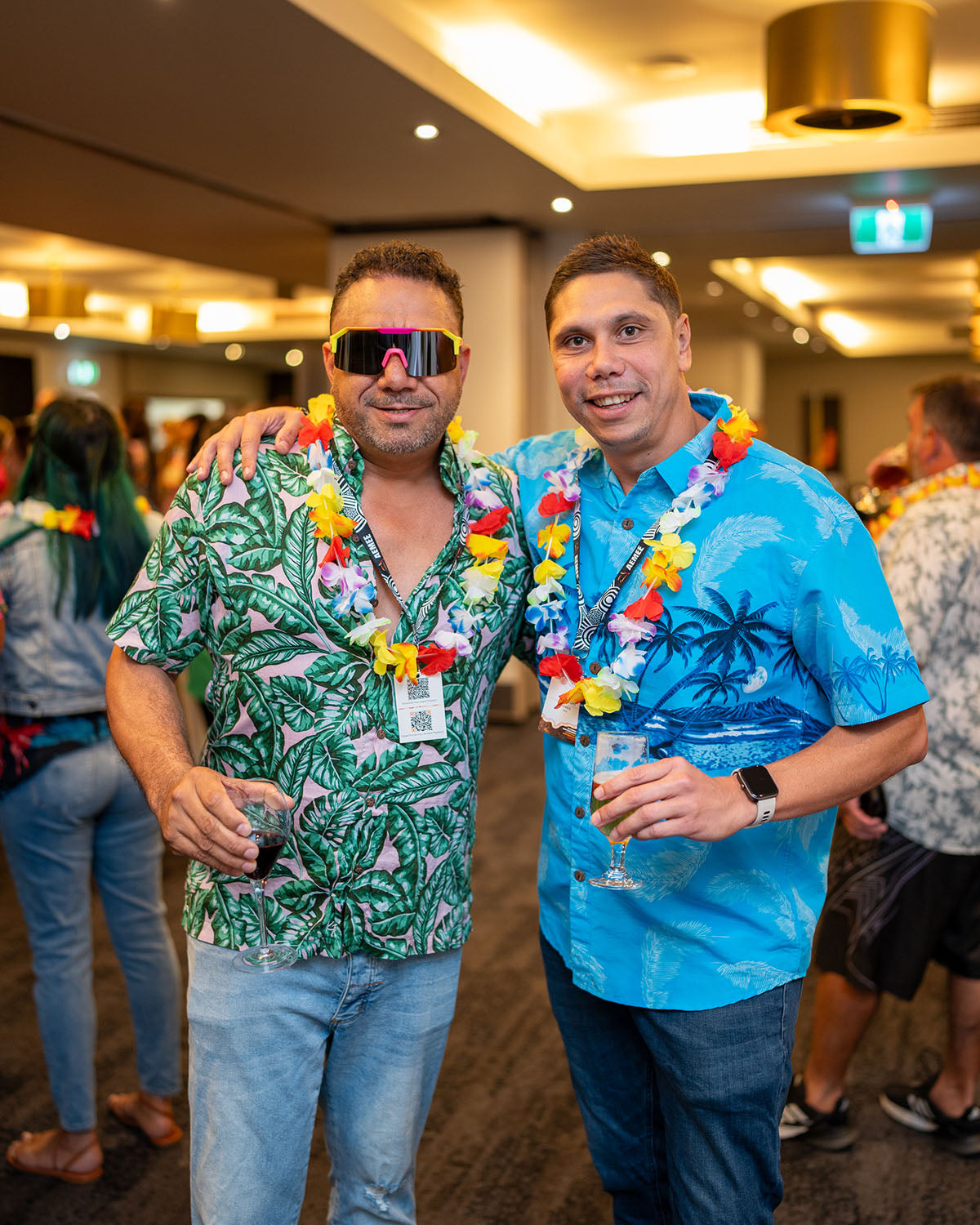 Two men wearing retro sunglasses, bright blue and green Hawaiian shirts, and decorative floral leis standing together at the evening mixer.