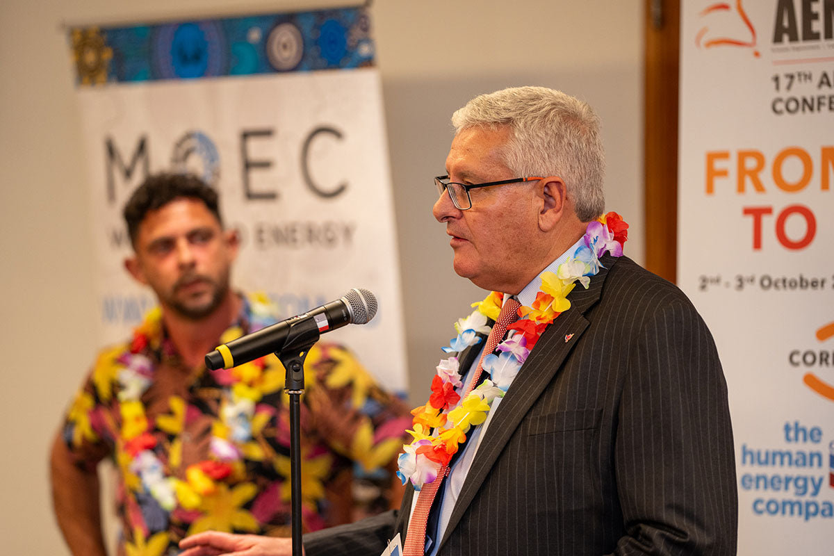 A senior corporate executive with a floral lei speaking into a microphone with the MOEC Water and Energy corporate partner banner visible behind him.