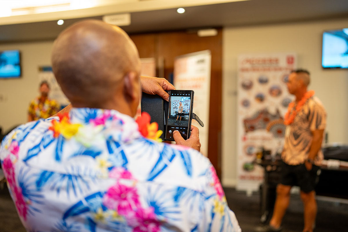 Over-the-shoulder view of a delegate in a bright floral shirt using his smartphone to take a photo of an attendee posing at the AEMEE social mixer.