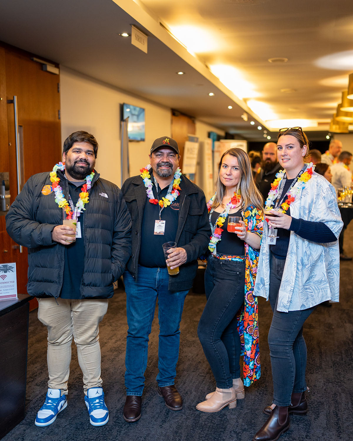 A group of four corporate partners and enterprise owners holding drinks and wearing colorful lei garlands while networking at the evening social function.