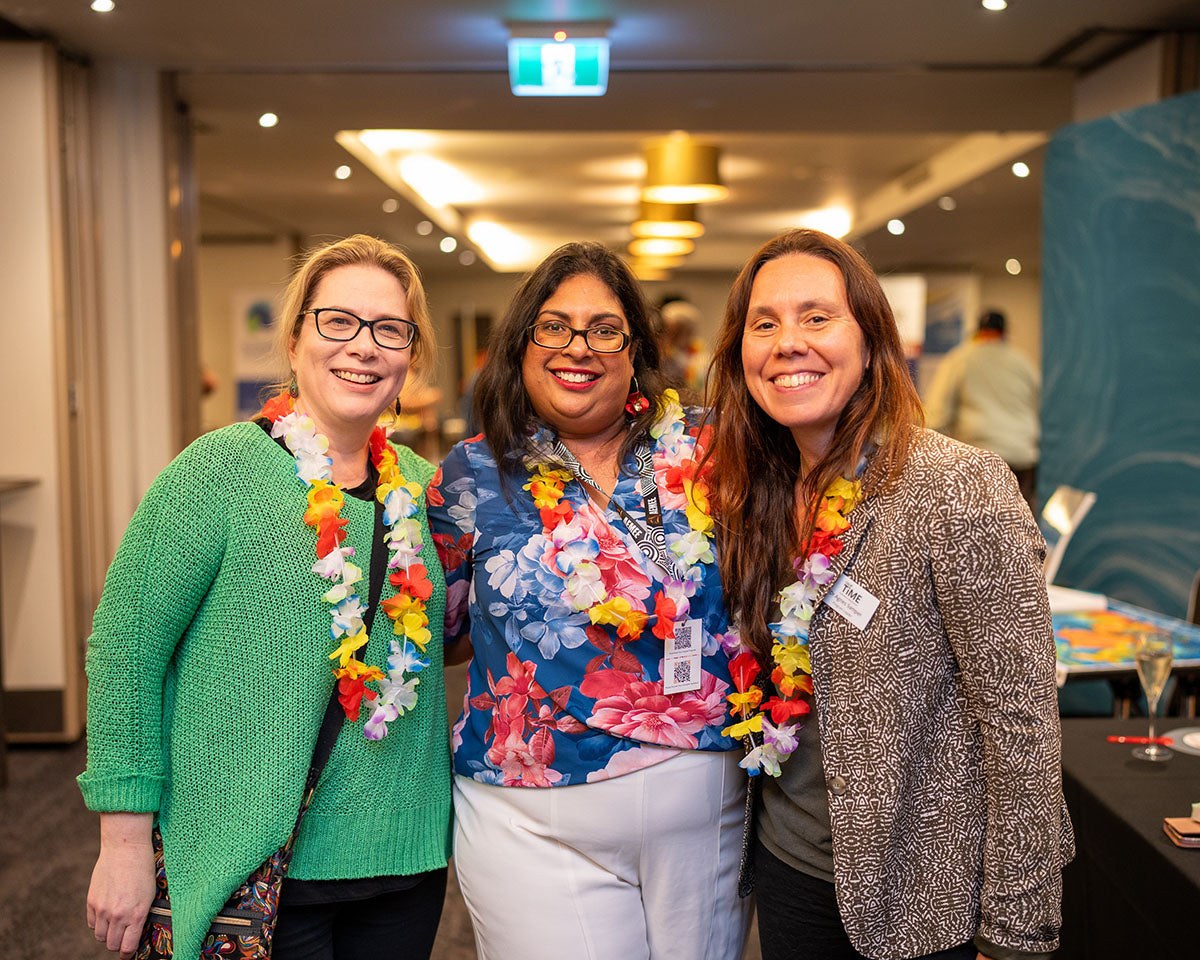 Three female industry professionals smiling together indoors, wearing decorative flower leis over casual conference attire at the annual corporate function.
