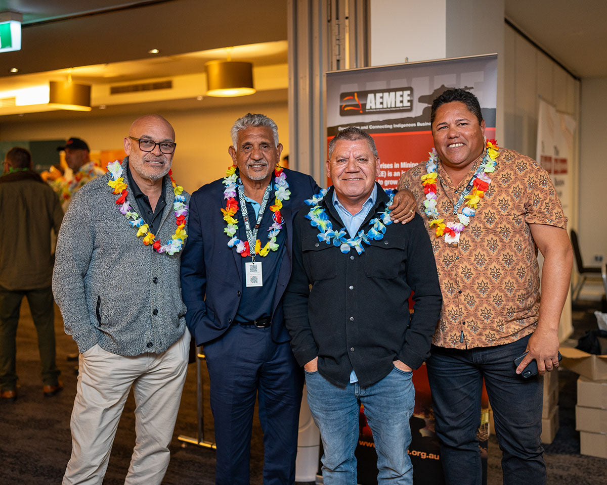 Four male industry executives wearing decorative flower leis smiling warmly together during the tropical-themed networking event.