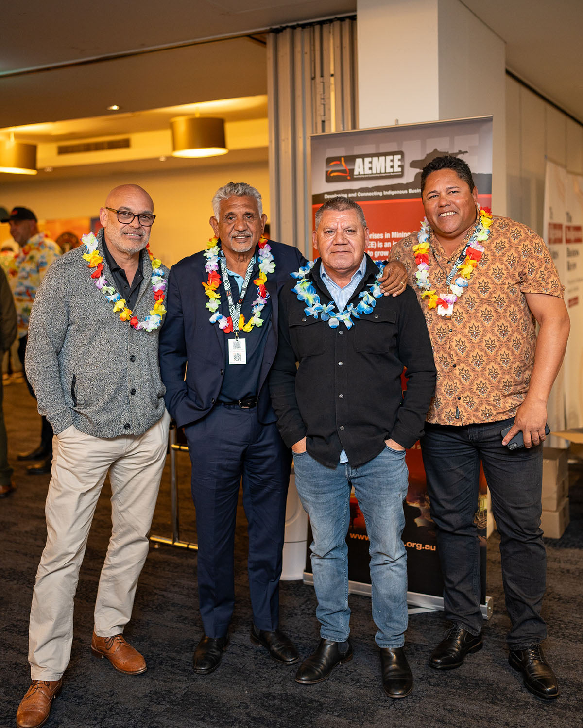 A group of four male delegates posing together with vibrant floral leis in front of an official AEMEE corporate partner display board.