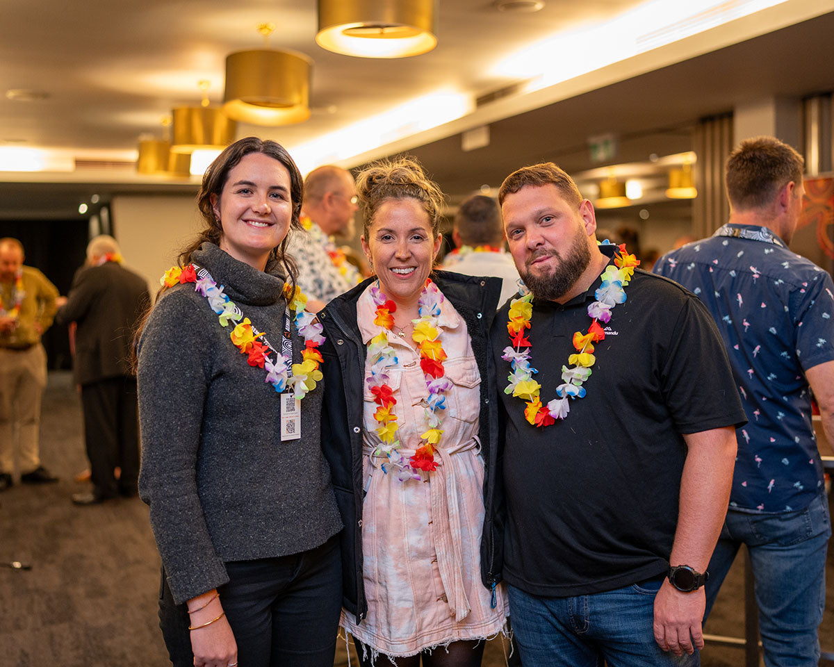 Two women and one man wearing matching flower leis smiling together inside the bustling function room at the 17th Annual AEMEE conference.
