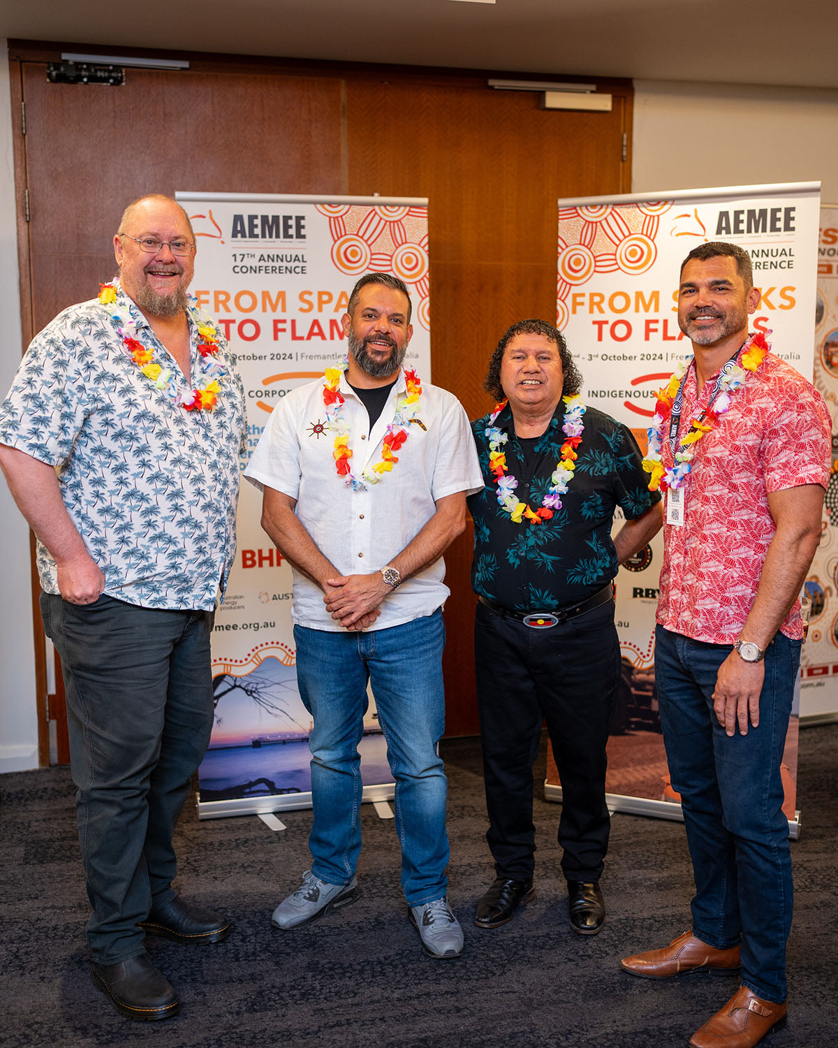 Four male attendees wearing Hawaiian resort shirts and flower leis standing together in front of the official "From Sparks to Flames" media banners.