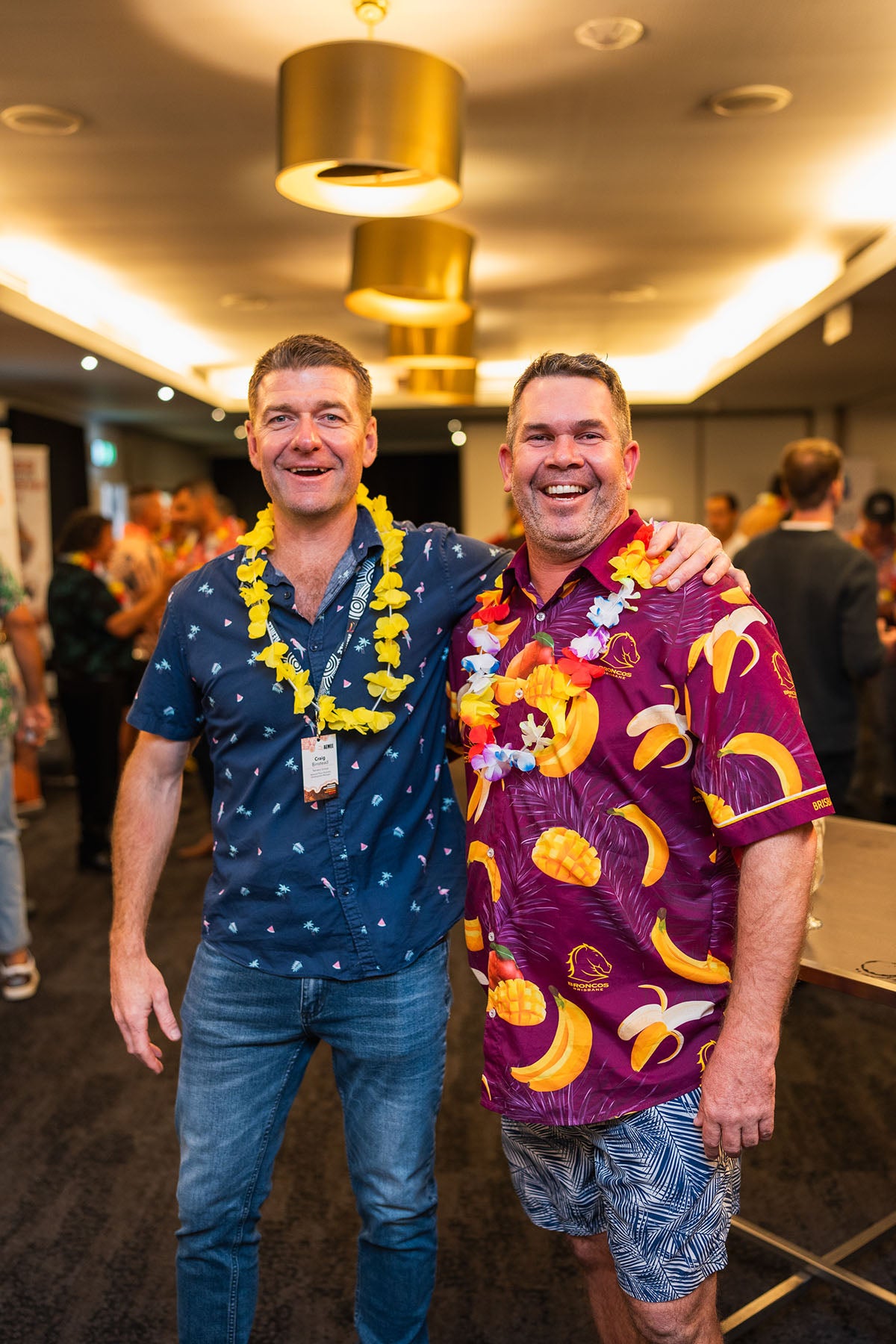 Two male corporate representatives with bright yellow flower leis laughing and embracing during the themed 2024 AEMEE conference networking celebration.