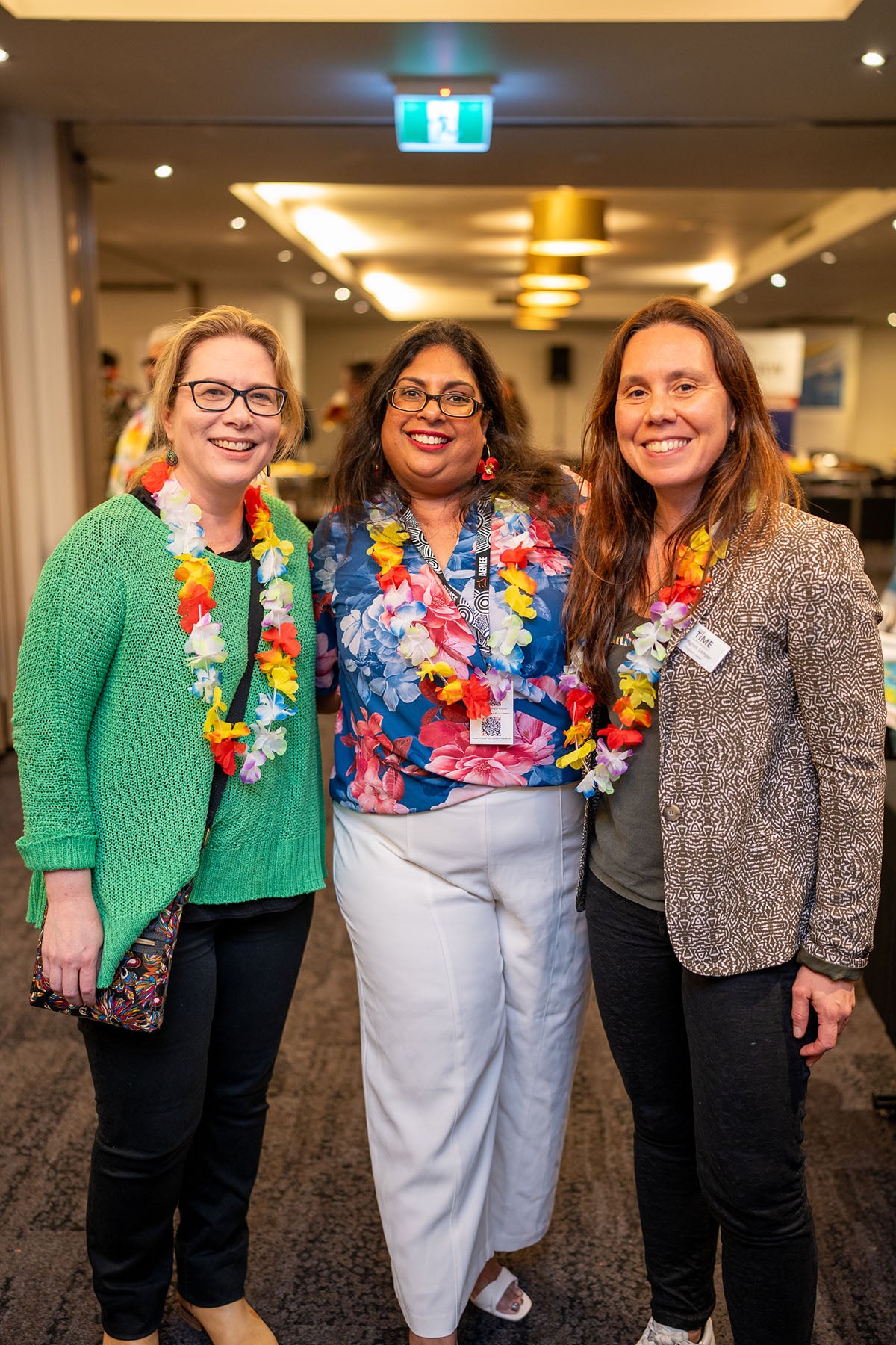 A medium group portrait of three female corporate partners wearing bright flower leis and smiling together during the tropical-themed networking evening.