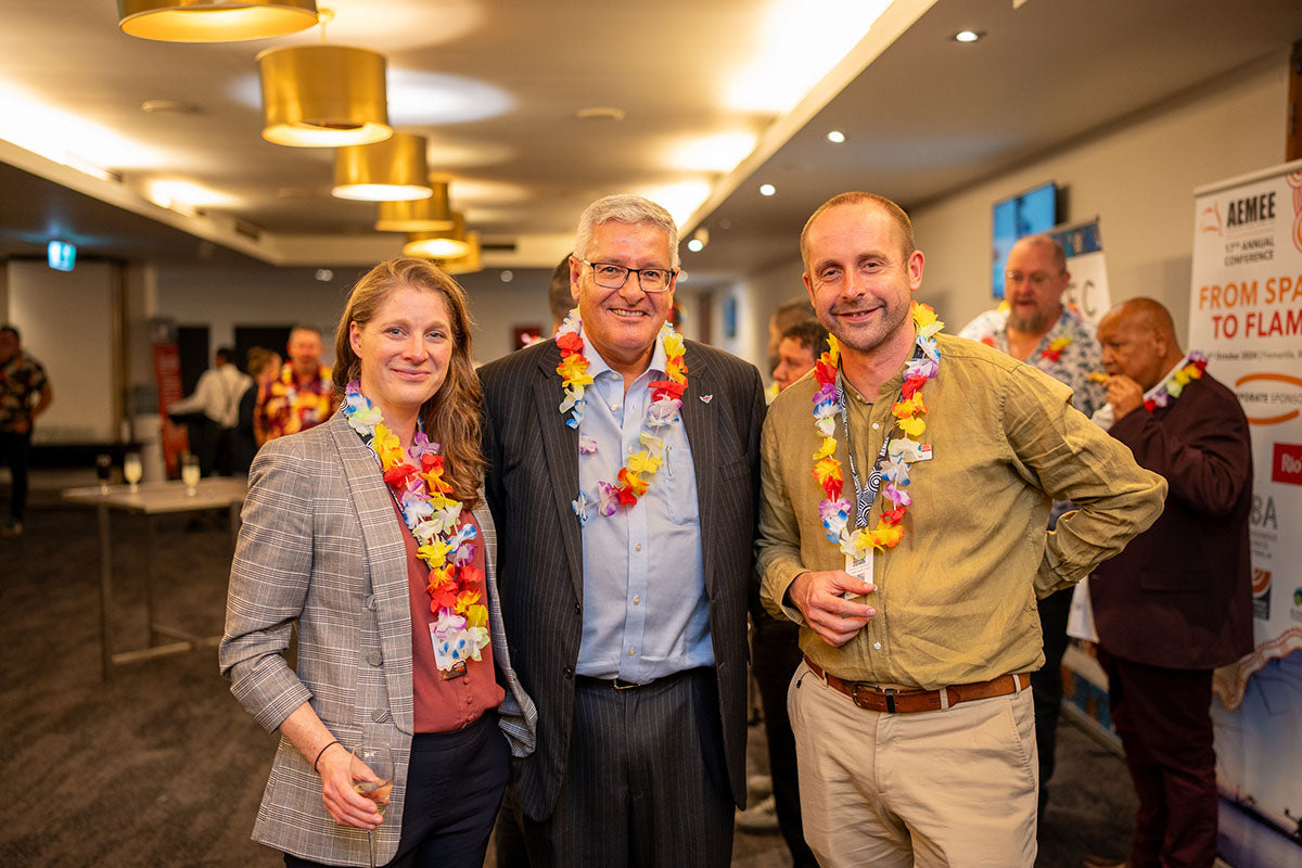 A group of three key event speaker, wearing festive tropical leis over their conference attire, smiling together inside the main venue hall.