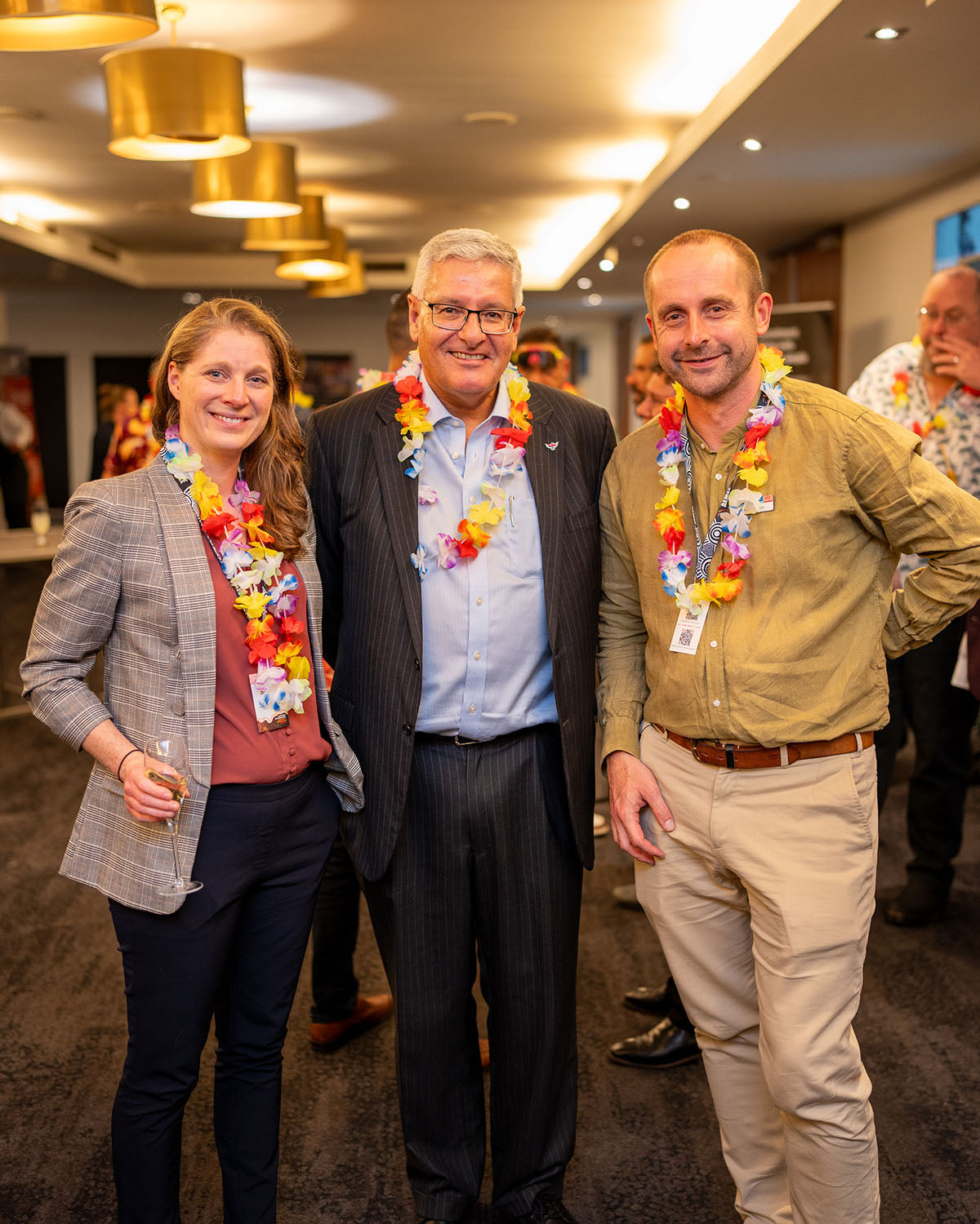 A medium group portrait of three industry professionals posing with colourful flower lei garlands during the themed corporate networking evening.