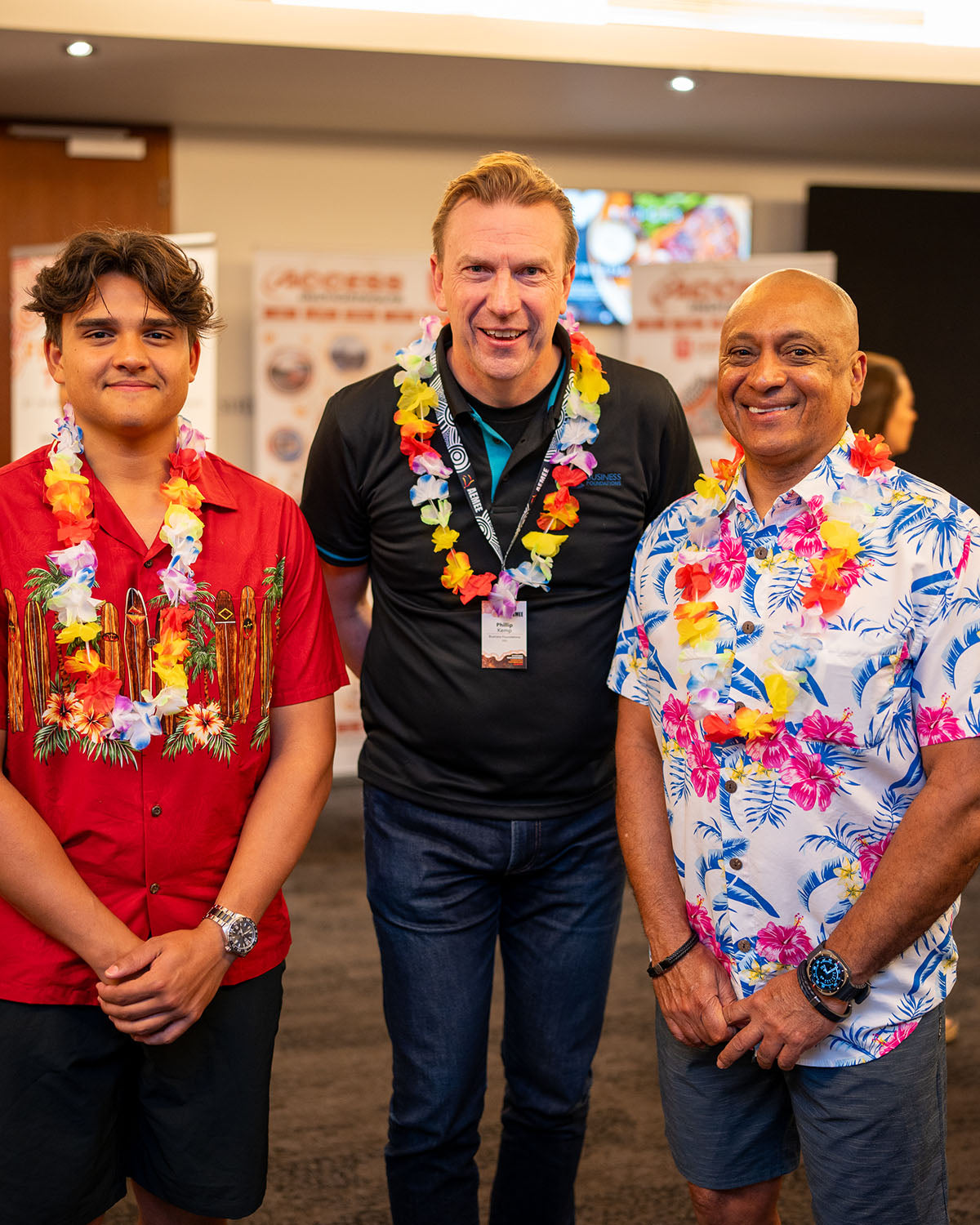 A centered portrait of three male delegates wearing colourful floral leis and resort-style shirts, smiling warmly inside the main event hall.
