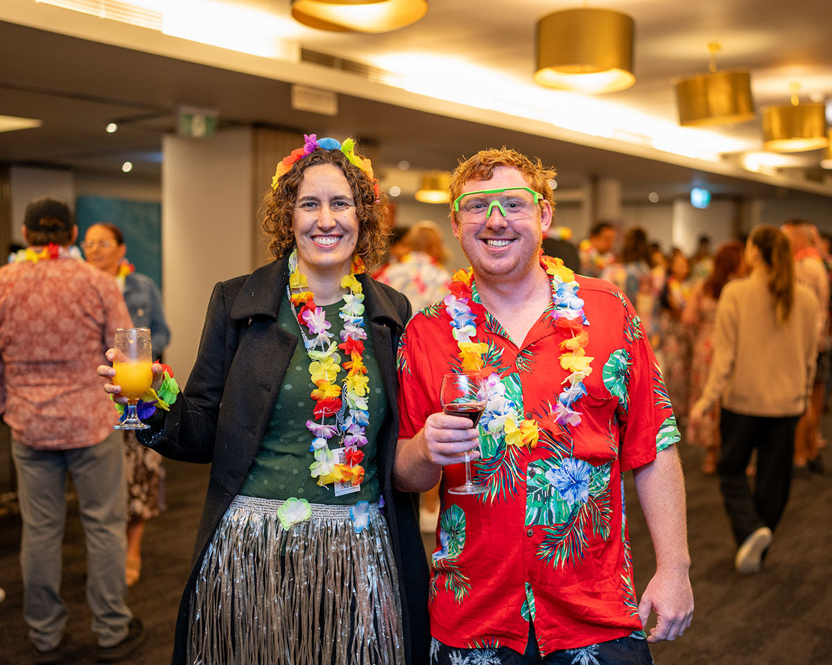Attendees wearing festive Hawaiian shirts, grass skirts, and neon glasses raising drinks and smiling during the 17th Annual AEMEE conference themed networking celebration.
