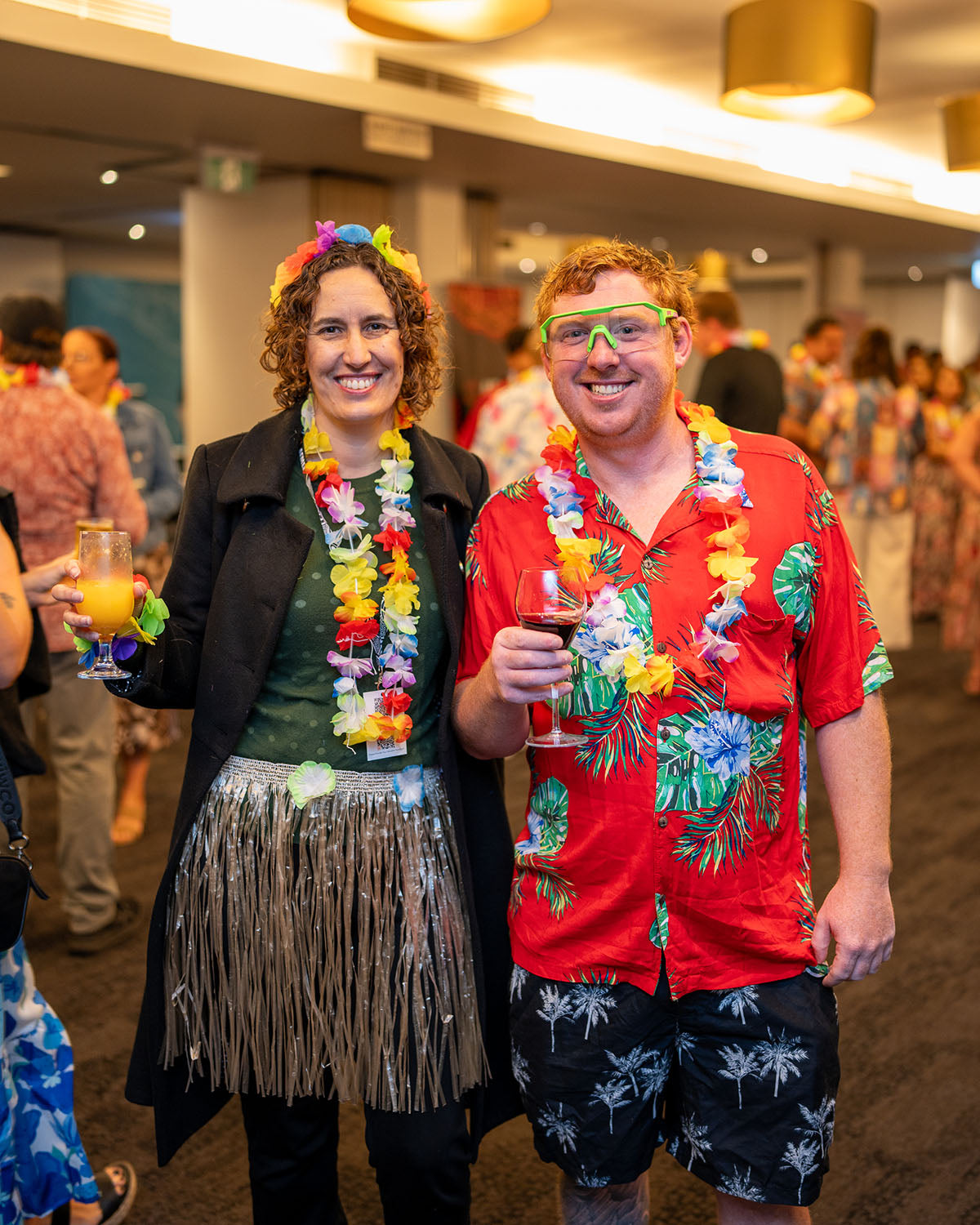 A male and female delegate posing together in fun island attire, including a grass skirt, neon green glasses, tropical shirts, and flower leis at the AEMEE social function.