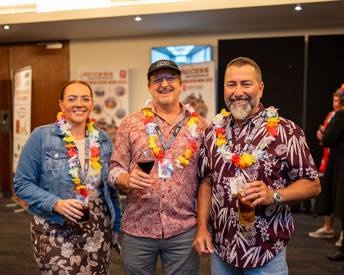 A group of three corporate delegates smiling and holding drinks while wearing vibrant floral leis at the 17th Annual AEMEE Networking Night.