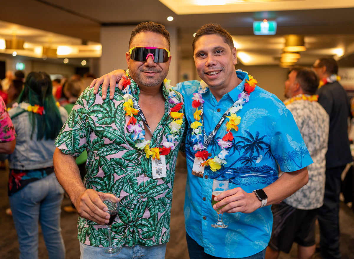 AEMEE CEO Jyi Lawton in his bright blue tropical palm shirt standing arm-in-arm with a fellow delegate wearing a pink banana-leaf shirt, neon sunglasses, and festive flower lei garlands.
