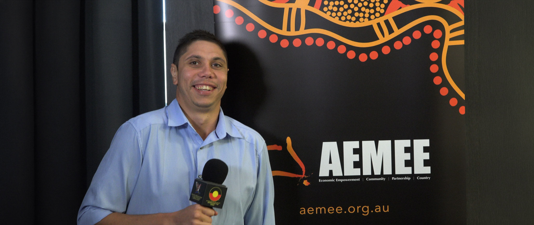 A historical 2019 photograph of a younger Jyi Lawton smiling broadly while holding a media microphone featuring the Aboriginal flag logo, standing in front of a black AEMEE pull-up banner adorned with red and orange traditional dot-art patterns.