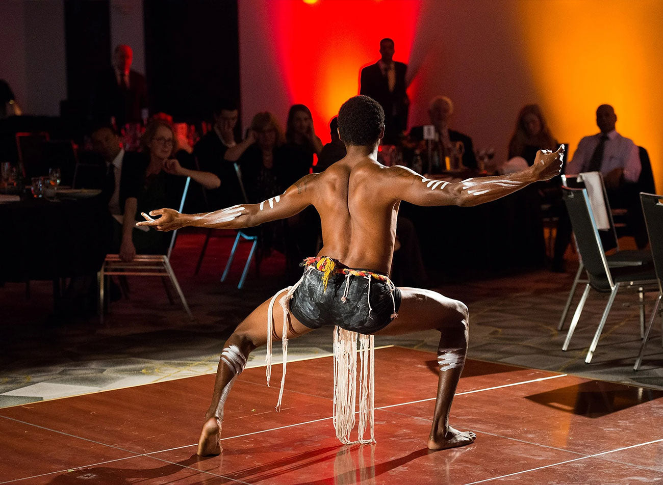 A Traditional First Nations dancer performing a ceremonial welcome on a wooden stage backdrop during the 2016 AEMEE National Conference in Canberra.