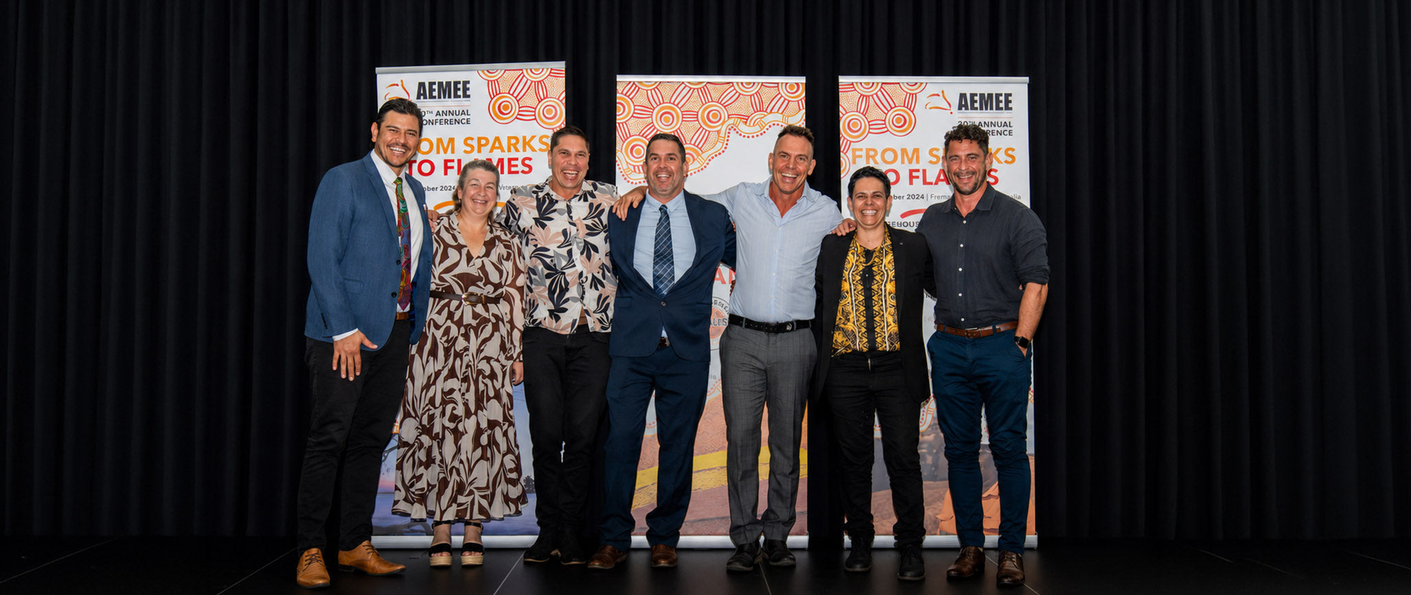 A group portrait of the AEMEE CEO and the Board of Directors standing together on stage in front of the official "From Sparks to Flames" media wall during the opening sequence of the gala night.