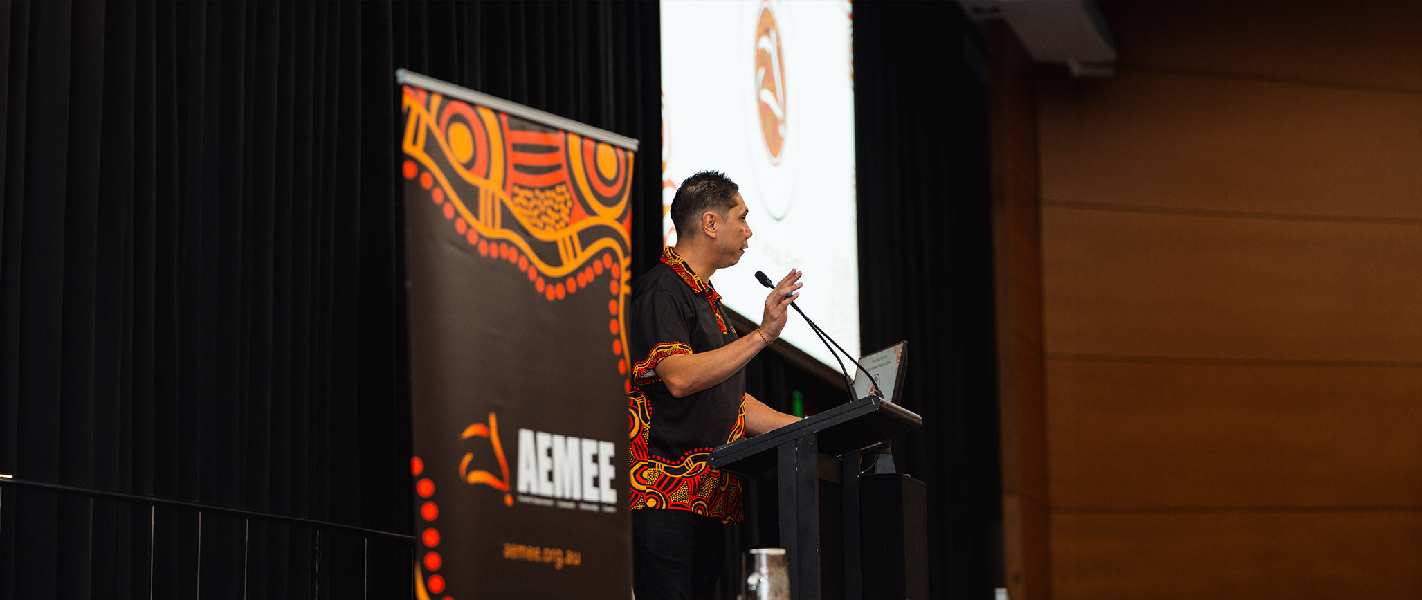 AEMEE CEO Jyi Lawton in profile, wearing a black shirt featuring vibrant orange and yellow traditional Indigenous patterns on the collar and hem, standing behind a dark wooden podium speaking into a microphone on stage next to an official AEMEE banner.