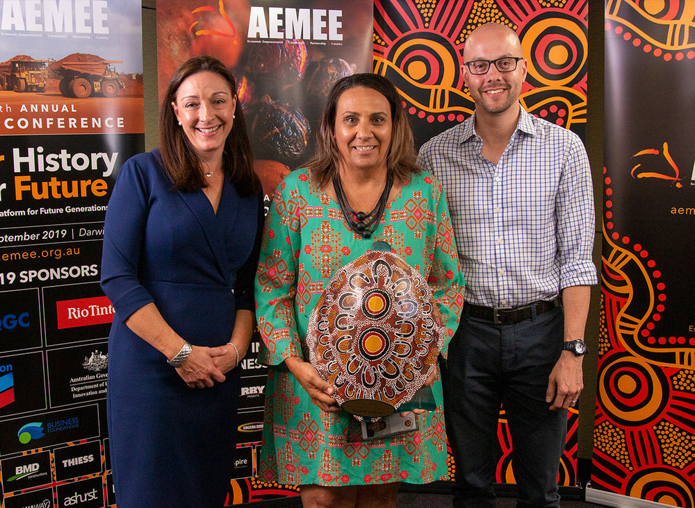 Three corporate professionals standing together on stage holding a hand-painted traditional shield trophy at the 2019 AEMEE conference awards ceremony in Darwin.
