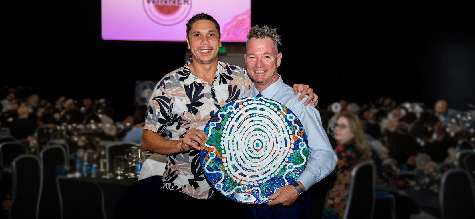 Two smiling corporate delegates holding a large hand-painted traditional circular shield trophy at an excellence awards gala, overlayed with introductory archive text for the AEMEE conference historical photo gallery.