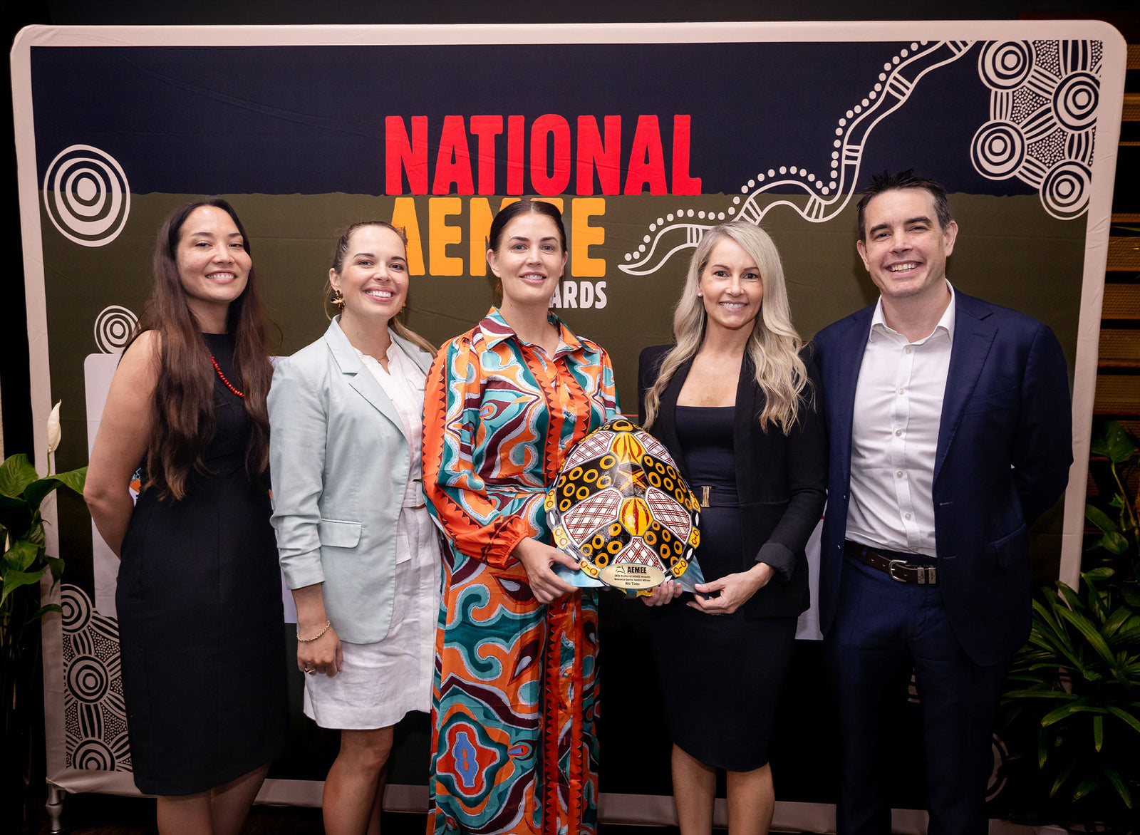 A diverse group of industry professionals and award winners holding a traditional shield trophy at the AEMEE Excellence Awards, representing success and community within the network.