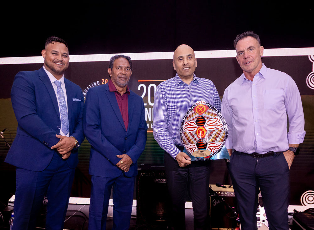 Four corporate executives in business attire smiling and standing together on stage while holding a hand-painted traditional shield award at an AEMEE gala event.