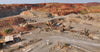 An expansive aerial drone photograph capturing the grand scale of an open-pit mining operation in the Australian outback, showcasing terraced excavation layers, deep red-ochre earth tones, and heavy industrial machinery contrasted against the vast natural landscape.